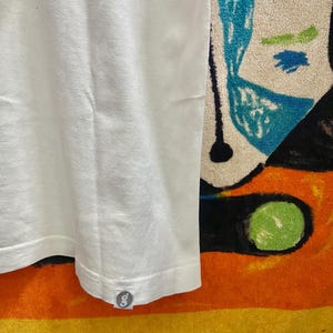 Puede incluir: Una camiseta blanca lisa con un peque&ntilde;o logotipo circular en el dobladillo. La tela tiene una apariencia texturizada. El fondo incluye un dise&ntilde;o abstracto colorido y la palabra "Picasso".