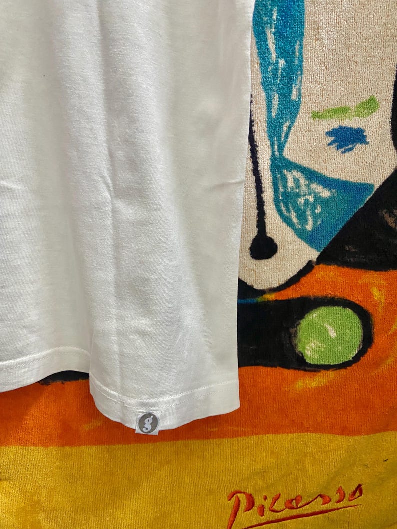 Puede incluir: Una camiseta blanca lisa con un peque&ntilde;o logotipo circular en el dobladillo. La tela tiene una apariencia texturizada. El fondo incluye un dise&ntilde;o abstracto colorido y la palabra "Picasso".