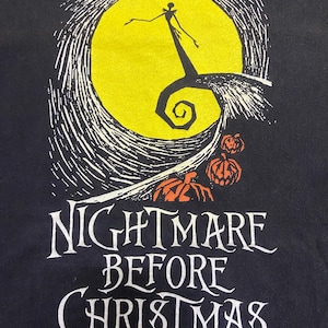 May include: Black t-shirt featuring a graphic from "The Nightmare Before Christmas." The design includes a yellow full moon, a silhouetted figure, and the words "Nightmare Before Christmas" in white lettering, with orange pumpkins.