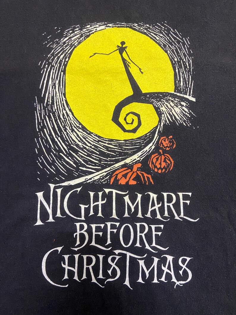May include: Black t-shirt featuring a graphic from "The Nightmare Before Christmas." The design includes a yellow full moon, a silhouetted figure, and the words "Nightmare Before Christmas" in white lettering, with orange pumpkins.