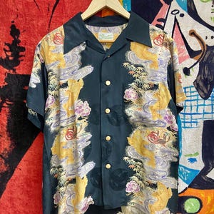 May include: A short-sleeved button-down shirt with a dark teal base color. The shirt features a repeating pattern of gold, purple, and white stylized imagery. The shirt has a camp collar and gold-colored buttons.