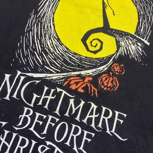 May include: Black t-shirt featuring a graphic from "The Nightmare Before Christmas." The design includes a yellow full moon, a silhouetted figure, and the text "NIGHTMARE BEFORE CHRISTMAS" in white, with orange pumpkins.