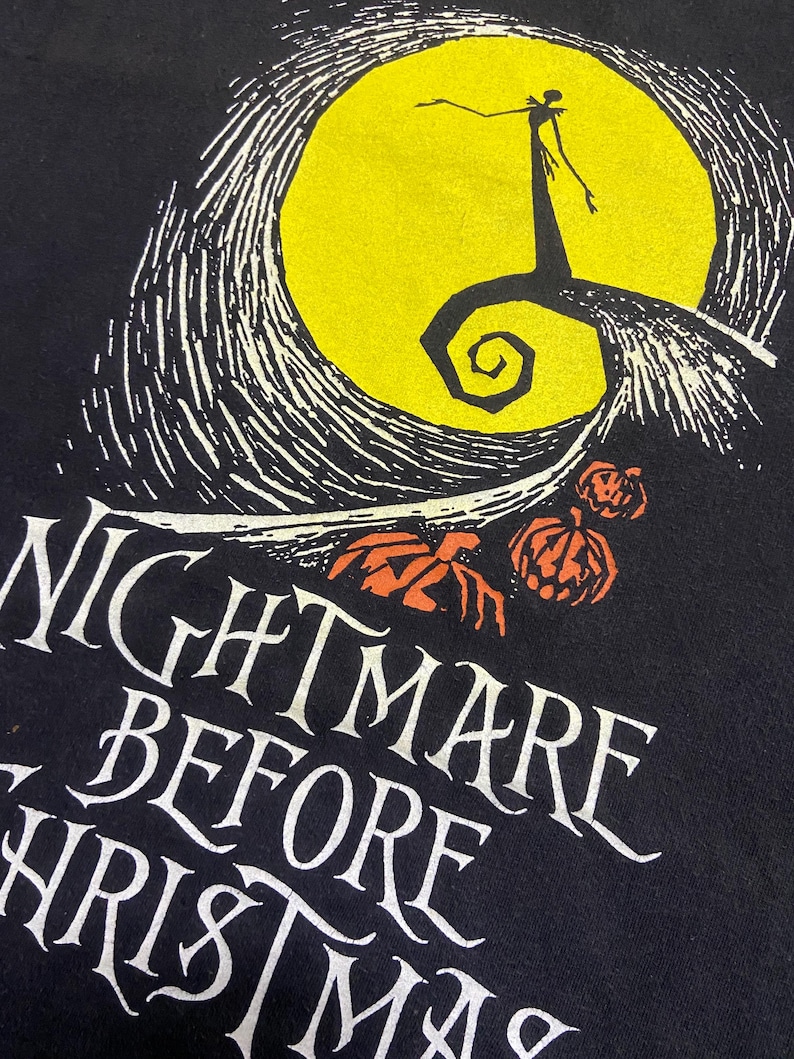 May include: Black t-shirt featuring a graphic from "The Nightmare Before Christmas." The design includes a yellow full moon, a silhouetted figure, and the text "NIGHTMARE BEFORE CHRISTMAS" in white, with orange pumpkins.