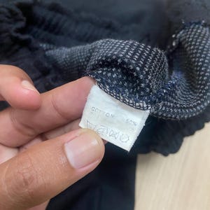 Puede incluir: Primer plano de tela negra con un patr&oacute;n texturizado y una etiqueta blanca. La etiqueta dice "COTTON 50%" con s&iacute;mbolos de lavado. La tela parece ser parte de una prenda, posiblemente pantalones cortos o largos.