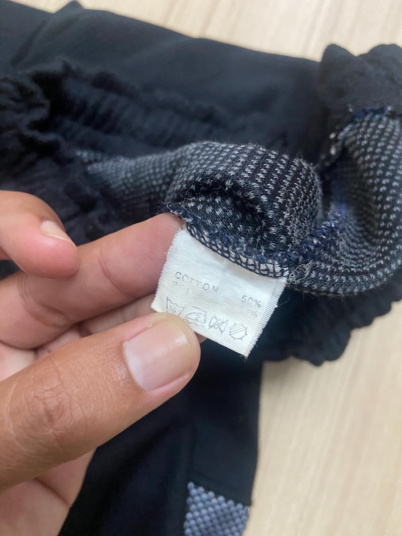 Puede incluir: Primer plano de tela negra con un patr&oacute;n texturizado y una etiqueta blanca. La etiqueta dice "COTTON 50%" con s&iacute;mbolos de lavado. La tela parece ser parte de una prenda, posiblemente pantalones cortos o largos.