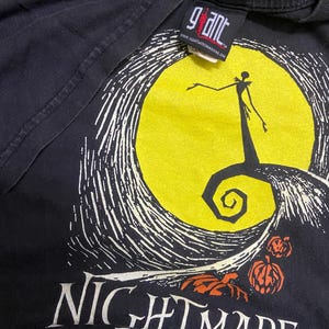 May include: Black t-shirt featuring a graphic from "The Nightmare Before Christmas." The design includes a yellow moon, a silhouette of Jack Skellington, and the film's title in white lettering. The shirt has a "Giant" tag.