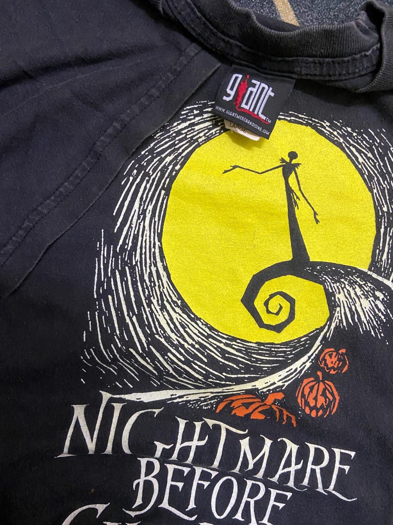 May include: Black t-shirt featuring a graphic from "The Nightmare Before Christmas." The design includes a yellow moon, a silhouette of Jack Skellington, and the film's title in white lettering. The shirt has a "Giant" tag.