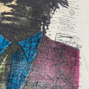 K&ouml;nnte beinhalten: Ein wei&szlig;es T-Shirt mit einem Schwarzwei&szlig;bild einer Person mit Afro. Das Bild ist mit bunten geometrischen Formen und Text &uuml;berlagert, darunter "BABAR THE TIGER", "MAGELLAN", "CIRCUS", "HAVANA", "BLUE RIBBON", "SERUM BOSA", "JOE LOUIS", "RADIUM", "ASIA", "EGYPT" und "DO".