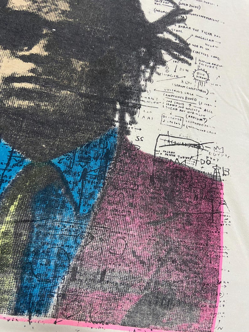 K&ouml;nnte beinhalten: Ein wei&szlig;es T-Shirt mit einem Schwarzwei&szlig;bild einer Person mit Afro. Das Bild ist mit bunten geometrischen Formen und Text &uuml;berlagert, darunter "BABAR THE TIGER", "MAGELLAN", "CIRCUS", "HAVANA", "BLUE RIBBON", "SERUM BOSA", "JOE LOUIS", "RADIUM", "ASIA", "EGYPT" und "DO".