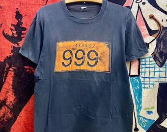 May include: A navy blue t-shirt with a rectangular orange graphic featuring the number "999" and the text "AS85.02". The shirt is hanging on a wooden hanger.