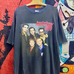 May include: Vintage black t-shirt featuring a group photo of the Backstreet Boys with the band's name in pink. The shirt has a dark blue collar and is hanging on a wooden hanger. The background is a colorful mix of abstract art.