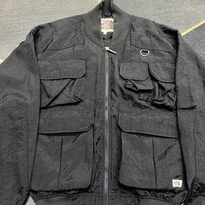 Peut inclure: Veste de bombardier noire avec plusieurs poches avant, une fermeture &eacute;clair et un col et des poignets c&ocirc;tel&eacute;s. La veste comporte un petit &eacute;cusson logo et un d&eacute;tail d'anneau m&eacute;tallique. Le logo The North Face est visible.