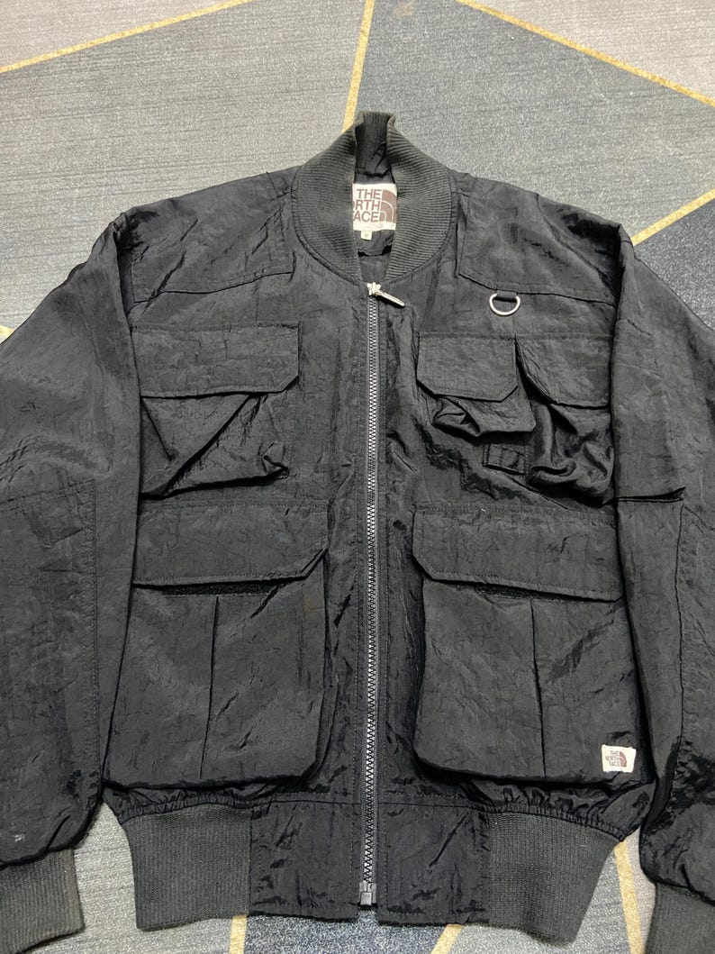 Peut inclure: Veste de bombardier noire avec plusieurs poches avant, une fermeture &eacute;clair et un col et des poignets c&ocirc;tel&eacute;s. La veste comporte un petit &eacute;cusson logo et un d&eacute;tail d'anneau m&eacute;tallique. Le logo The North Face est visible.