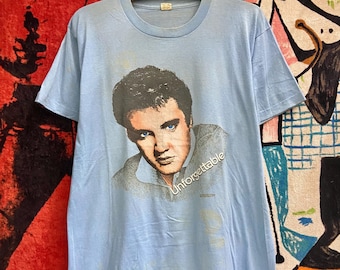 May include: Light blue t-shirt with a vintage image of Elvis Presley. The shirt has the word "Unforgettable" printed on it.