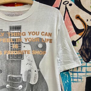 Puede incluir: Camiseta blanca con un dise&ntilde;o de guitarra en blanco y negro y el texto "The only thing you can feel in your life is favorite shop." Hecha de algod&oacute;n.