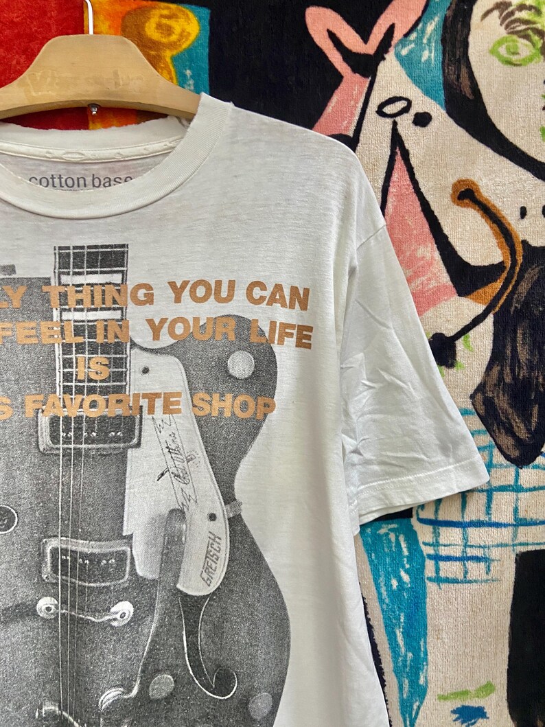 Puede incluir: Camiseta blanca con un dise&ntilde;o de guitarra en blanco y negro y el texto "The only thing you can feel in your life is favorite shop." Hecha de algod&oacute;n.