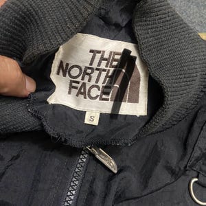 Peut inclure: Veste noire avec un col c&ocirc;tel&eacute; gris et une fermeture &eacute;clair. La veste comporte une &eacute;tiquette blanche avec "THE NORTH FACE" imprim&eacute; en noir et marron. L'&eacute;tiquette porte &eacute;galement une petite &eacute;tiquette de taille "S".