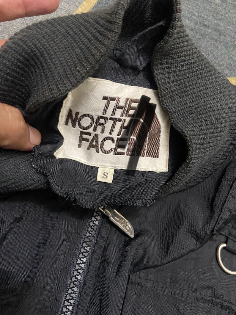 Peut inclure: Veste noire avec un col c&ocirc;tel&eacute; gris et une fermeture &eacute;clair. La veste comporte une &eacute;tiquette blanche avec "THE NORTH FACE" imprim&eacute; en noir et marron. L'&eacute;tiquette porte &eacute;galement une petite &eacute;tiquette de taille "S".