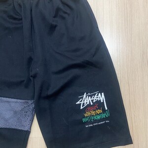Puede incluir: Pantalones cortos deportivos negros con un panel de malla blanca y azul en la pierna izquierda. La pierna derecha presenta el logotipo de Stussy y texto en blanco, verde, amarillo y rojo. Los pantalones cortos tienen cintura el&aacute;stica.