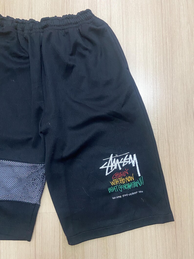 Puede incluir: Pantalones cortos deportivos negros con un panel de malla blanca y azul en la pierna izquierda. La pierna derecha presenta el logotipo de Stussy y texto en blanco, verde, amarillo y rojo. Los pantalones cortos tienen cintura el&aacute;stica.