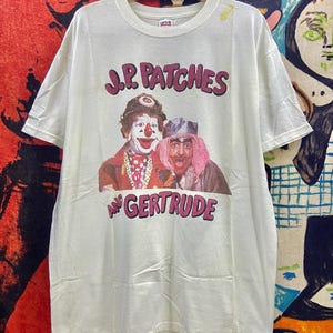 May include: Cream-coloured t-shirt with a graphic of two clowns and the text "J.P. Patches and Gertrude" in maroon. The clowns are in colourful costumes. The shirt is displayed on a wooden hanger.