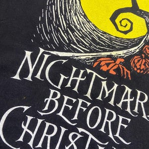 May include: Black t-shirt featuring the words "Nightmare Before Christmas" in white, stylized lettering. The design includes a yellow moon, a silhouetted tree, and orange pumpkins.