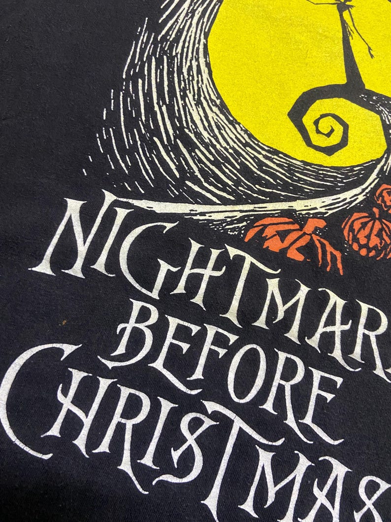 May include: Black t-shirt featuring the words "Nightmare Before Christmas" in white, stylized lettering. The design includes a yellow moon, a silhouetted tree, and orange pumpkins.