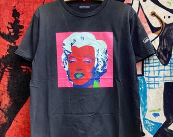 May include: Black t-shirt featuring a graphic print of Marilyn Monroe's face in a pop art style. The graphic is in pink, red, blue, and green. The shirt has a black tag with the word "mastermind" printed on it.
