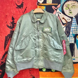 May include: Sage green Alpha Industries bomber jacket with a zip closure, two front pockets, and a small patch on the chest. The jacket has a ribbed hem and cuffs. A red "REMOVE BEFORE FLIGHT" tag hangs from the sleeve.