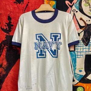 May include: Cream-colored vintage t-shirt with navy blue trim around the collar and sleeves. The shirt features the word "NAVY" in large, block letters in blue. The shirt is hanging on a wooden hanger.