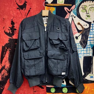 Peut inclure: Veste de bombardier noire avec plusieurs poches avant et une fermeture &eacute;clair. La veste a un col, des poignets et un ourlet c&ocirc;tel&eacute;s. Le logo The North Face est visible sur la poitrine. La veste est suspendue &agrave; un cintre en bois.