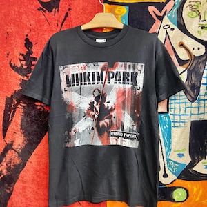 Könnte beinhalten: Schwarzes T-Shirt mit dem Bandnamen "Linkin Park" in weißen Blockbuchstaben über einer Grafik einer Figur mit rotem und weißem Hintergrund. Der Text "Hybrid Theory" steht darunter. Das T-Shirt hängt an einem Holzbügel.