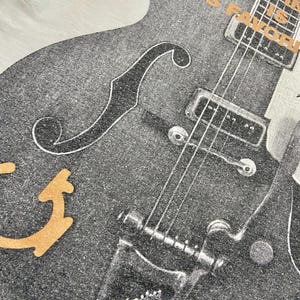 Puede incluir: Primer plano de una camiseta blanca con un gr&aacute;fico de guitarra en blanco y negro. El dise&ntilde;o de la guitarra incluye cuerdas, un puente y una efe. El texto "NICO'S FAVORITE" est&aacute; impreso en naranja encima de la guitarra.