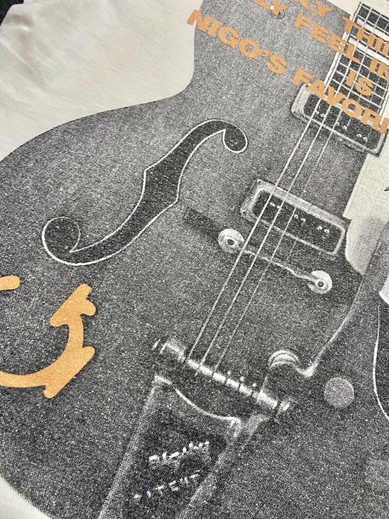 Puede incluir: Primer plano de una camiseta blanca con un gr&aacute;fico de guitarra en blanco y negro. El dise&ntilde;o de la guitarra incluye cuerdas, un puente y una efe. El texto "NICO'S FAVORITE" est&aacute; impreso en naranja encima de la guitarra.