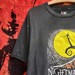 May include: Black t-shirt featuring a graphic from "The Nightmare Before Christmas." The design includes a yellow moon, Jack Skellington, and the film's title in white lettering. The shirt is hanging on a wooden hanger.
