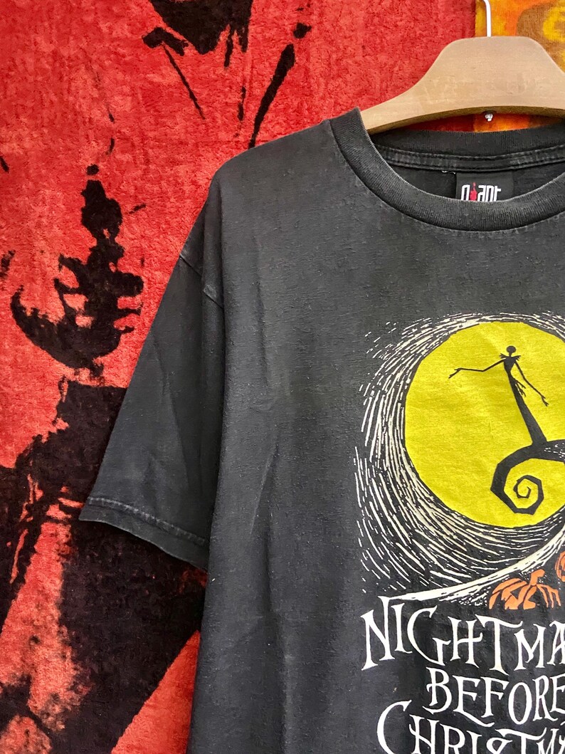 May include: Black t-shirt featuring a graphic from "The Nightmare Before Christmas." The design includes a yellow moon, Jack Skellington, and the film's title in white lettering. The shirt is hanging on a wooden hanger.