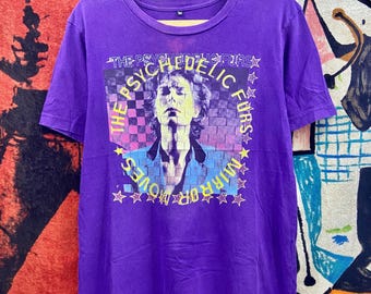 May include: A purple t-shirt featuring a graphic of The Psychedelic Furs band. The design includes a portrait of a person with the words "The Psychedelic Furs" and "Mirror Moves" in a star-studded design. The shirt is hanging on a wooden hanger.