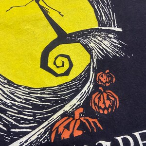 May include: Black t-shirt featuring a yellow full moon with a silhouetted figure and a spiral design. Orange carved pumpkins are below the moon. The word "NIGHTMARE" is visible at the bottom.