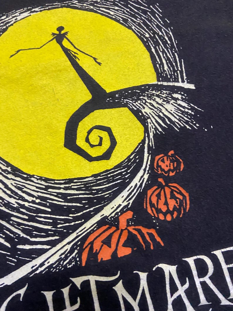 May include: Black t-shirt featuring a yellow full moon with a silhouetted figure and a spiral design. Orange carved pumpkins are below the moon. The word "NIGHTMARE" is visible at the bottom.