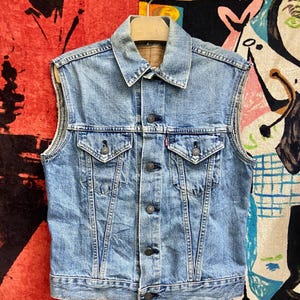 May include: A light blue denim vest featuring a classic collar, a button-down front closure, and two chest pockets with buttoned flaps. The garment has a faded wash, visible stitching details, and a small red tab on the left chest pocket.