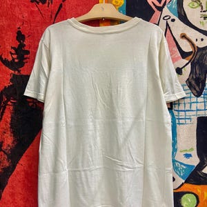 K&ouml;nnte beinhalten: Ein wei&szlig;es T-Shirt mit kurzen &Auml;rmeln und einem Rundhalsausschnitt. Das Shirt h&auml;ngt an einem Holzb&uuml;gel vor einem bunten Hintergrund.