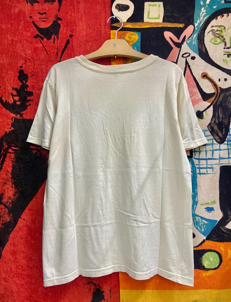 K&ouml;nnte beinhalten: Ein wei&szlig;es T-Shirt mit kurzen &Auml;rmeln und einem Rundhalsausschnitt. Das Shirt h&auml;ngt an einem Holzb&uuml;gel vor einem bunten Hintergrund.