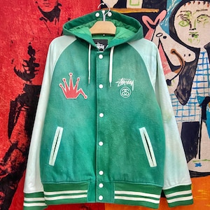 May include: Green and white Stussy varsity jacket with a hood. The jacket features a red crown patch and the Stussy logo in white. It has white sleeves, white buttons, and green and white striped trim at the waist and cuffs.