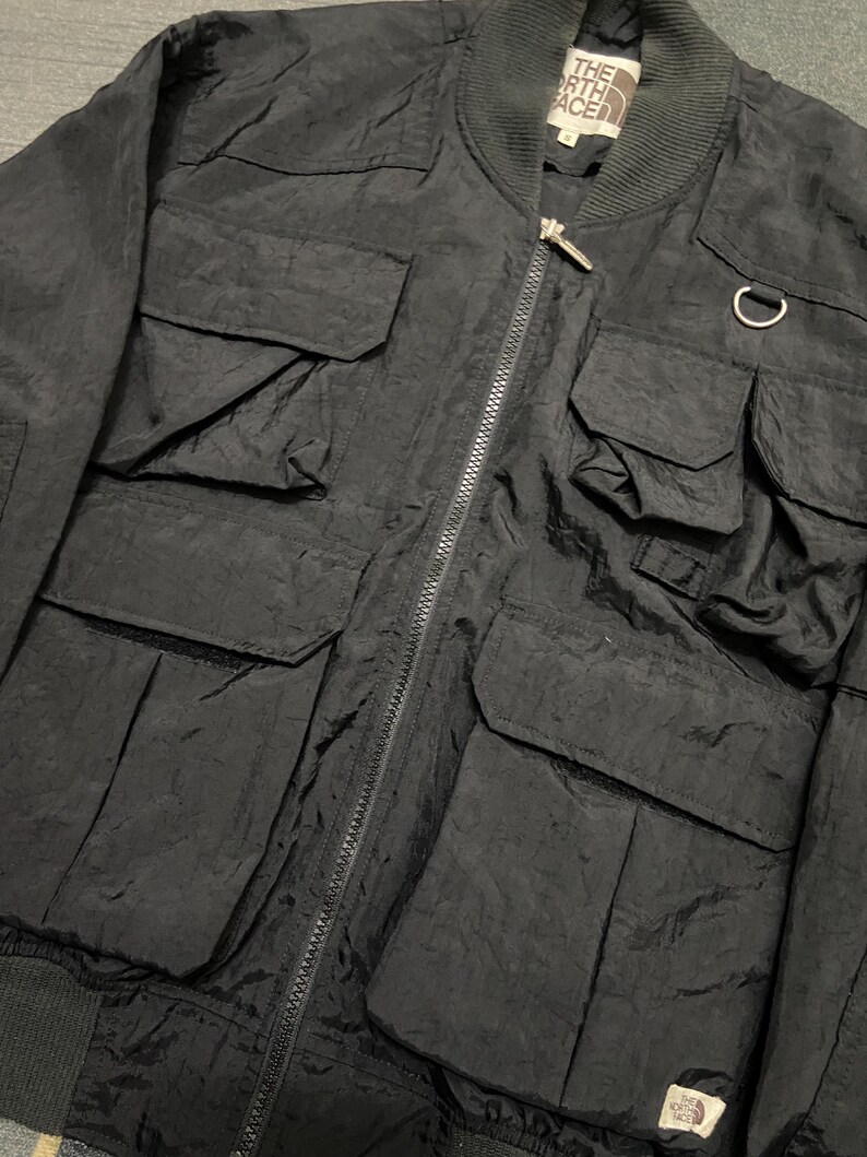 Peut inclure: Veste de bombardier noire avec plusieurs poches avant et une fermeture &eacute;clair. La veste pr&eacute;sente un col et des poignets c&ocirc;tel&eacute;s, et un petit &eacute;cusson logo. Le logo The North Face est visible.