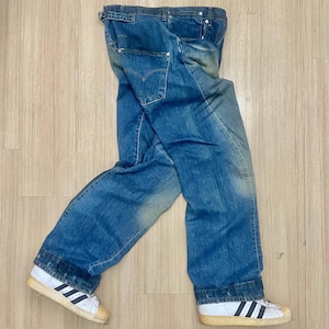 Puede incluir: Vaqueros azules descoloridos con un diseño clásico de cinco bolsillos. Los vaqueros se combinan con zapatillas blancas con rayas negras y suela color canela. Los vaqueros están colocados sobre una superficie de madera marrón claro.