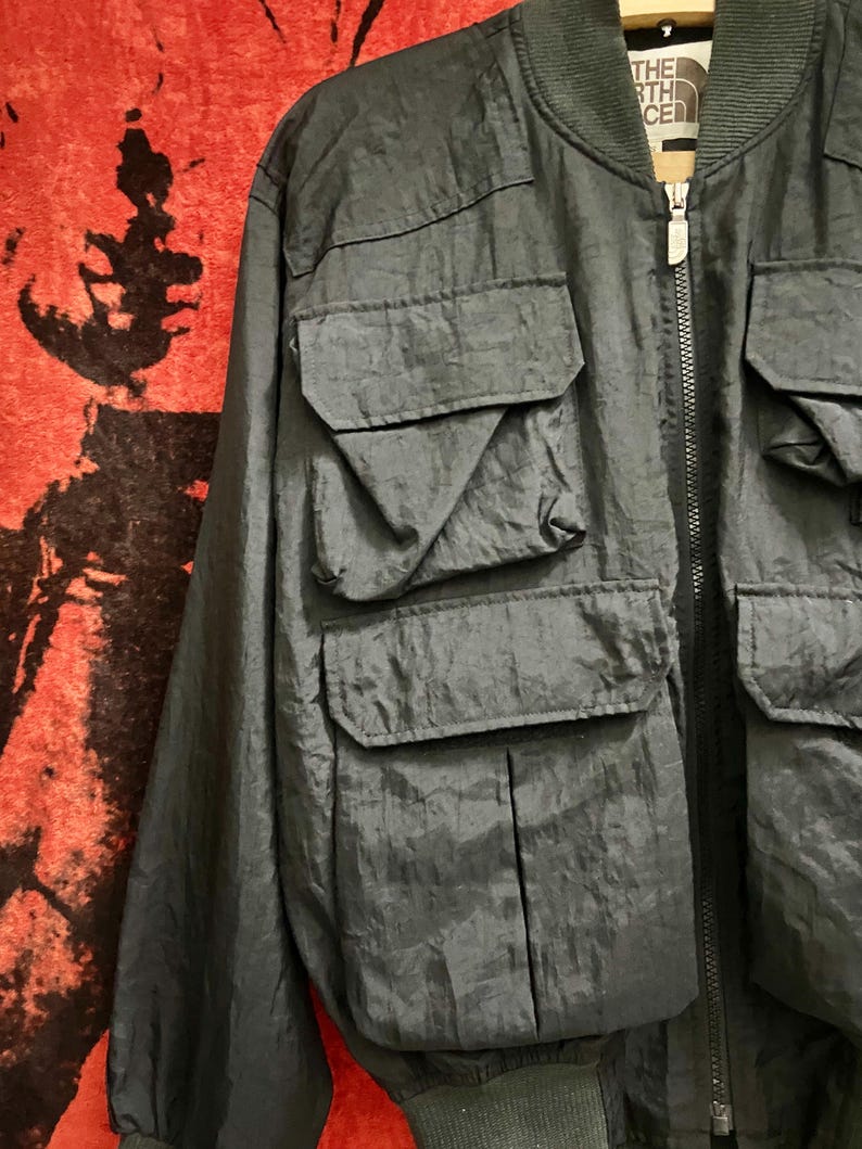 Peut inclure: Veste de bombardier noire avec plusieurs poches avant et une fermeture &eacute;clair. La veste porte le nom de la marque "The North Face" sur le col. La veste a un col et des poignets c&ocirc;tel&eacute;s. Le tissu est l&eacute;g&egrave;rement brillant.