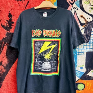 以下が含まれることがあります： 赤と黄色の文字で「BAD BRAINS」と書かれたグラフィックプリントが施された黒の半袖Tシャツ。デザインには、白い建物のドームに落ちる黄色い稲妻が描かれており、赤、黄、緑の枠で囲まれています。