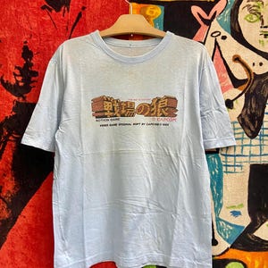 May include: A pale blue t-shirt featuring a graphic from the video game "Commando". The graphic includes Japanese text and the Capcom logo. The shirt is suspended from a wooden hanger.