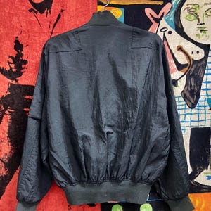 Peut inclure: Veste de bombardier bleu marine fonc&eacute; avec col, poignets et ourlet c&ocirc;tel&eacute;s. La veste a une texture brillante et l&eacute;g&egrave;rement froiss&eacute;e et une fermeture &eacute;clair sur toute la longueur. La veste est suspendue &agrave; un cintre blanc sur un fond color&eacute;.