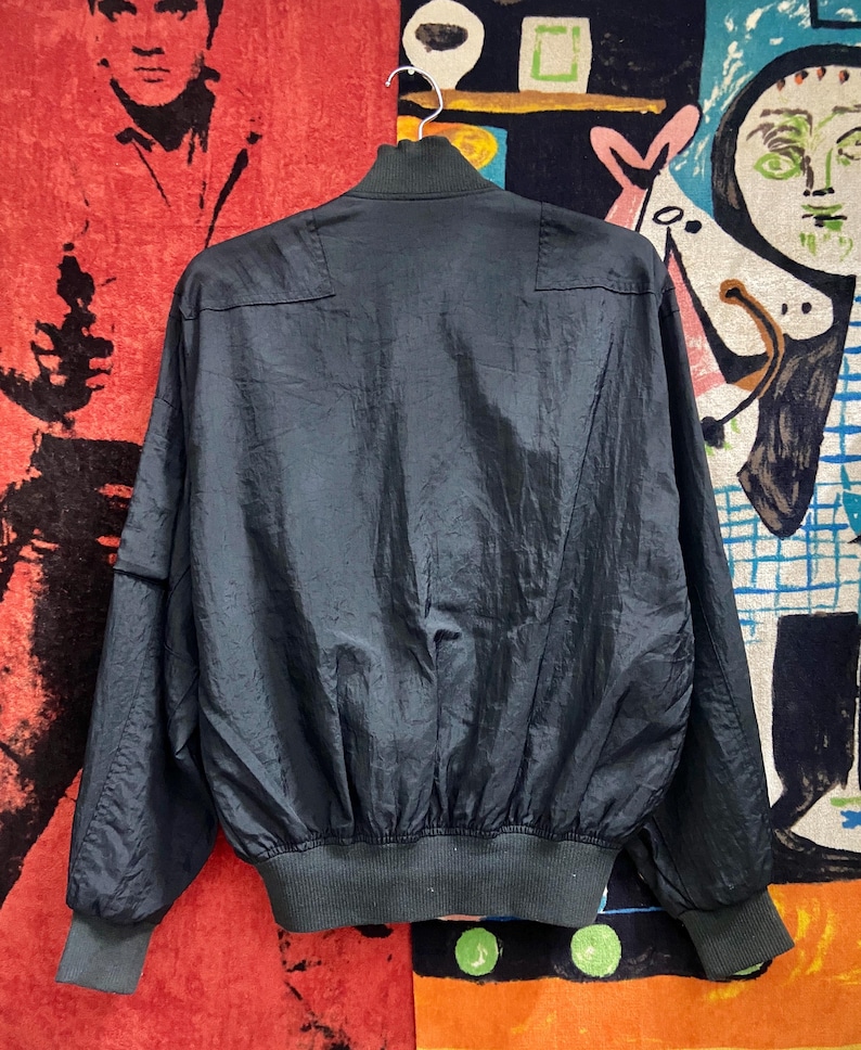 Peut inclure: Veste de bombardier bleu marine fonc&eacute; avec col, poignets et ourlet c&ocirc;tel&eacute;s. La veste a une texture brillante et l&eacute;g&egrave;rement froiss&eacute;e et une fermeture &eacute;clair sur toute la longueur. La veste est suspendue &agrave; un cintre blanc sur un fond color&eacute;.